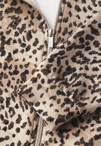 Gros plan sur un tissu imprimé léopard montrant un motif beige et noir avec une fermeture éclair en argent et des détails de manches à revers.