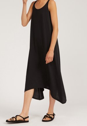 Kobieta w czarnej, bezrękawnikowej sukience midi z asymetrycznym dołem i czarnych sandałach z plecionymi paskami, stojąca na tle jednolitego tła.