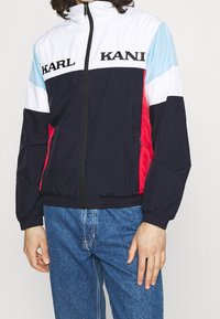 Väriblokkipilttuinen tuulitakki laivastonsinisellä, valkoisella ja vaaleansinisellä, jossa on vetoketju, joustavat hihansuut ja rinnan kohdalla kirjailtu "KARL"-logo.