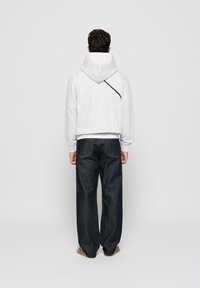 Lys grå hettegenser med snor og asymmetrisk svart aksent, kombinert med løse mørke denimbukser, sett fra baksiden.