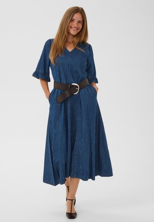Femme aux longs cheveux portant une robe midi en denim bleu avec des manches arrivant aux coudes et une large ceinture noire, souriante avec les mains dans les poches.