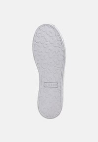 Valkoinen tennarin pohja, jossa on rakenteellisia, abstrakteja kuvioita ja "GUESS"-logo kohokirjoitettuna keskelle.