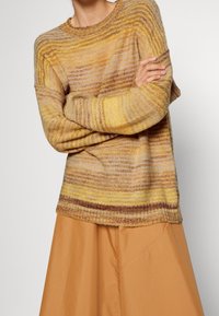 Pull en maille avec des rayures horizontales en tons jaunes, beiges et bruns. Encolure arrondie et coupe ample, associé à une jupe beige.