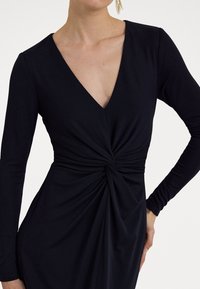 Svart långärmad klänning med djup V-ringning och snurrad knut vid midjan, med en mjuk och slät textur. Åtsittande silhuett.