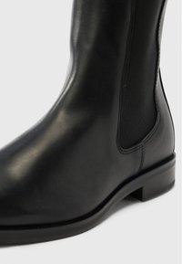 Černá kotníková bota z hladké kůže s elastickými bočními panely, zaoblenou špičkou a nízkým blokovým podpatkem. Má jemné šicí detaily.