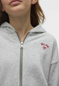 Grå zip-up hoodie i en mjuk bomullsblandning, med en framzipper, stor huva och röd broderad text med en rosettdetalj på bröstet.