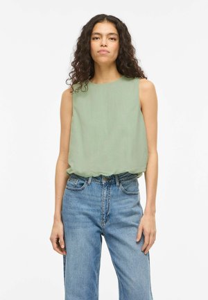 Mladá žena s dlhými kučeravými vlasmi, oblečená v bezrukávovej svetlozelenej blúzke a vysoko pásových modrých džínsoch, stojaca na bielom pozadí.