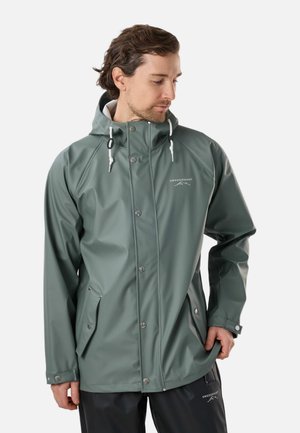 Mann trägt grüne wasserdichte Jacke mit Kapuze und schwarze Hose, schaut nach unten und hält mit einer Hand den Saum der Jacke.