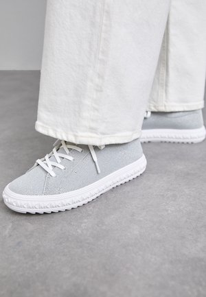 Lysegrå mesh-sneakers med hvide snørebånd båret med hvide bukser med vide ben på en grå gulv.