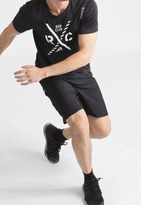 Musta urheilupaita, jossa on "RUN CLUB" -graafinen kuvaus, sekä mustat shortsit. Malli, joka käyttää mustia juoksukenkiä ja mustia sukkia, aktiivisessa asennossa.