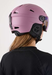 Rosa Skihelm mit matter Oberfläche, verstellbarem Visier, Belüftungsschlitzen und schwarzen Akzenten. Befestigte Gurte sorgen für Sicherheit unter dem Kinn.