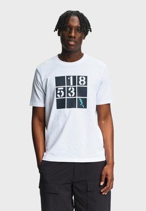 Homme aux dreadlocks courtes portant un T-shirt blanc avec un motif en grille numérotée noir et turquoise et un pantalon cargo noir, debout.