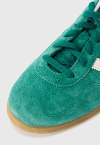 Sapatilha de camurça azul-petróleo com superfície texturizada, apresentando biqueira redonda, fecho de cordões e sola em borracha natural. Detalhes brancos na lateral.