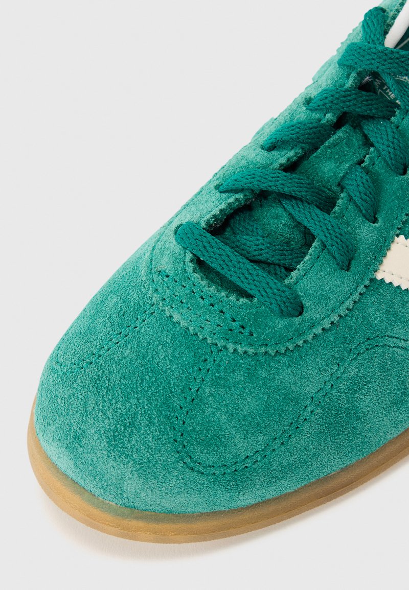 Teal ruskind sneakers med fremtrædende syninger, rund tå, flad gummisål og kontrasterende hvide striber på siden. Snørebåndene er teal.