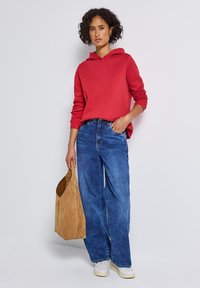 Femme aux cheveux bouclés portant un sweat à capuche rouge, un jean bleu à pattes d'éléphant, des baskets blanches, tenant un sac fourre-tout en daim beige, adossée à un mur blanc.