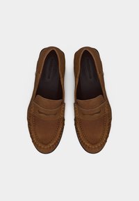 Καφέ μοκασίνια σουέντ με ραμμένα σχέδια και λουράκια penny, φτιαγμένα από πάνω σε λευκό απλό φόντο.