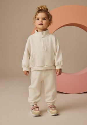 Sweatshirt en polaire texturée couleur crème avec fermeture éclair demi-longueur et poche avant, associé à un jogger assorti et des baskets multicolores.