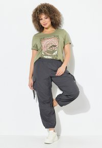 Olijfgroene t-shirt met grafische print, gecombineerd met donkergrijze cargobroek met zijzakken, en witte sneakers met een plateauzool.