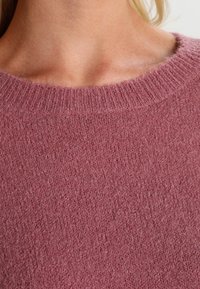 Maglione rosa lavorato a maglia con scollo girocollo a costine e una texture soffice e pelosa, che evidenzia un aspetto caldo e accogliente.