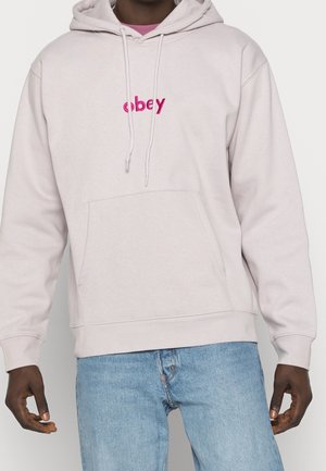 Personne portant un sweat à capuche beige clair avec un texte rose "obey" sur la poitrine et un jean bleu clair, debout, les bras relâchés sur les côtés.