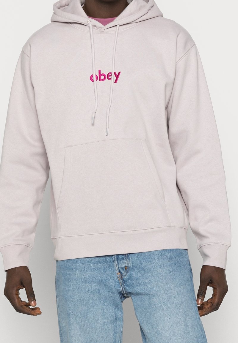 Personne portant un sweat à capuche beige clair avec un texte rose "obey" sur la poitrine et un jean bleu clair, debout, les bras relâchés sur les côtés.