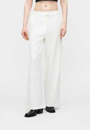 Femme portant un pantalon blanc évasé à taille haute et des bottines noires, debout devant un fond clair uni.