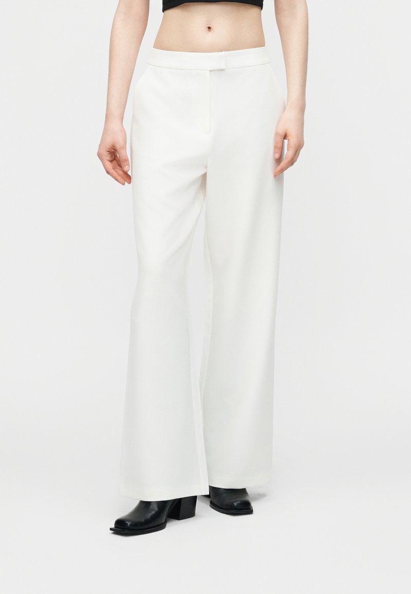 Femme portant un pantalon blanc évasé à taille haute et des bottines noires, debout devant un fond clair uni.