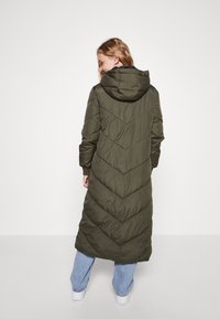 Casaco puffer verde-oliva com capuz, apresentando um padrão acolchoado em forma de chevron, de comprimento longo e mangas com punhos, usado sobre jeans de tom azul claro.