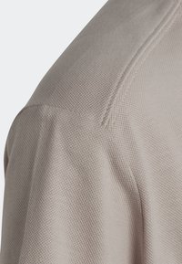 Lichtbeige textiel met structuur, close-up van de schouder en naaddetails van het kledingstuk, waarbij het geweven patroon en de zachte val worden benadrukt.