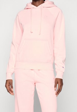 Personne portant un sweat à capuche rose clair et un pantalon de survêtement assorti avec poche avant et cordons, debout devant un fond uni.