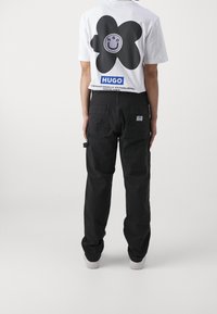 Weißes T-Shirt mit grafischem schwarzen Blumenmuster und Logo auf der Rückseite, kombiniert mit schwarzen Cargo-Hosen mit hinteren Taschen und seitlichen Schlaufen.