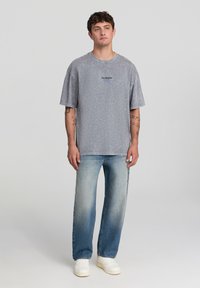 Un giovane con i capelli corti indossa una t-shirt oversize grigia sbiadita con la scritta "Los Angeles", jeans azzurri chiari, scarpe da ginnastica bianche e tatuaggi visibili sulle braccia.