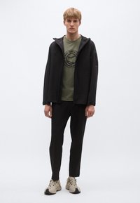 Fekete kapucnis dzseki, olívazöld póló köralakú logóval, fekete nadrág és bézs sportcipő texturált fűzőkkel és részletekkel.