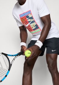 Camiseta blanca con un diseño gráfico colorido que presenta patrones de animales. Pantalones cortos negros. Sosteniendo una raqueta de tenis y una pelota de tenis amarilla.