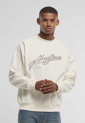 Młody mężczyzna w białej bluzie z logo „Kani Jeans”, stoi ze złączonymi rękami na gładkim szarym tle.
