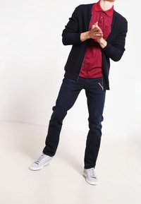 Mörk marinblå cardigan över en vinröd polotröja, i kombination med mörka jeans och vita sneakers. Släta texturer och figursydd design genomgående.