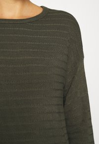 Gros plan d'une personne portant un pull en maille côtelée vert olive avec un col rond, sur un fond uni clair.