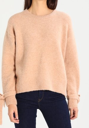 Lichtroze trui van zacht, pluizig materiaal, met een ronde hals, geribbelde manchetten en zoom, en strikken aan de mouwen. Gemaakt in combinatie met donkere jeans.