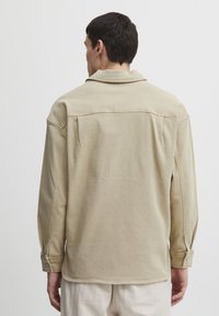 Beige denim jacket with a soft texture, buttoned cuffs, and a collar. Features a yoke design and visible stitching details.