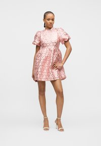 Γυναίκα με κοτσίδες, ντυμένη με κοντό, γυαλιστερό ροζ φόρεμα με φουσκωτά μανίκια και λευκά ψηλοτάκουνα σανδάλια, στέκεται με το χέρι στη μέση μπροστά σε λευκό φόντο.