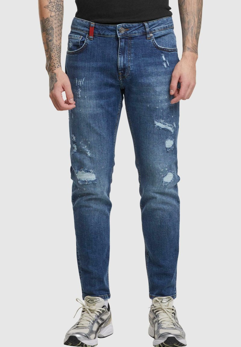 Jean en denim bleu délavé avec des zones déchirées, coupe slim, et style à cinq poches. Porté avec des baskets grises et argentées.