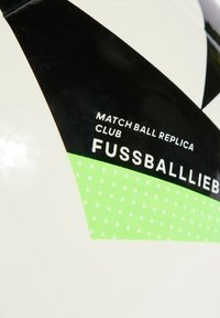 Bílý fotbalový míč s černými a zelenými akcenty. Má texturovaný povrch a tištěný text: "REPLIKA ZÁVODNÍHO MÍČE KLUB FUSSBALLLIEBE."