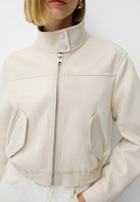 Cream-colored jacket made of smooth fabric, featuring a high collar, front zipper, and two side pockets with curved edges. Elasticized hem.