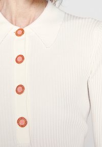 Haut blanc à côtes avec un col classique et cinq boutons orange au centre rose, orné de petits strass transparents.