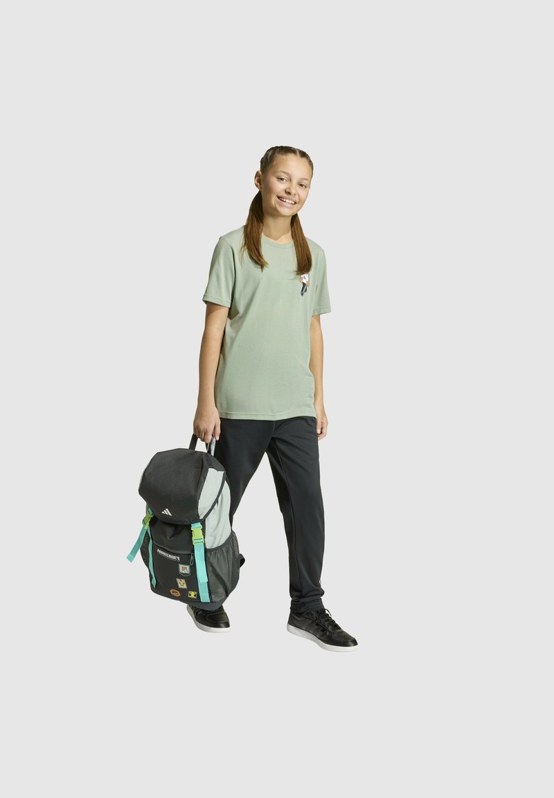 Sivi i svijetlozeleni ruksak s crnim detaljima, podesivim remenima i zakrpama. Model nosi svijetlozeleno majicu i crne hlače.