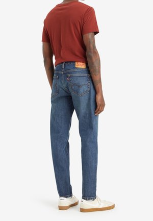 Osoba ubrana w niebieskie dżinsy Levi's oraz białe trampki z gumowymi podeszwami, stojąca tyłem, w rdzawej koszulce z krótkim rękawem.