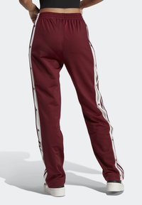 Pantalones deportivos burdeos con rayas laterales blancas y botones a presión. Cintura elástica, ajuste tapered, fabricados con tejido suave. Detalles de marca mínimos.