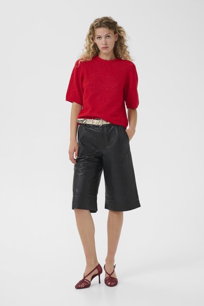 Femme aux cheveux blonds bouclés portant un pull rouge à manches courtes, un short en cuir noir longueur genou, une ceinture à motif serpent et des sandales à talons rouges.