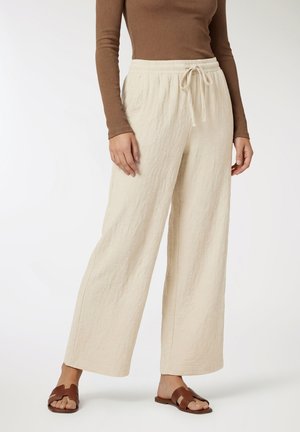 Kobieta w beżowych szerokich spodniach z elastycznym pasem i sznurkiem, brązowym topie z długim rękawem oraz brązowych sandałach na otwartym palcu, stojąca na białym tle.