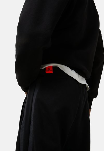 Sort sweatshirt med en tekstureret overflade og et rødt mærketag, båret over hvidt stof. Sorte bukser med sidelister komplementerer outfittet.